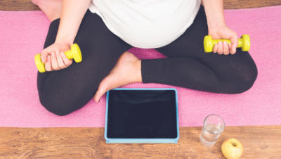 Bird's eye view of pregnant woman preparing to do yoga from iPad, holding weights, with apple and glass of water by her side